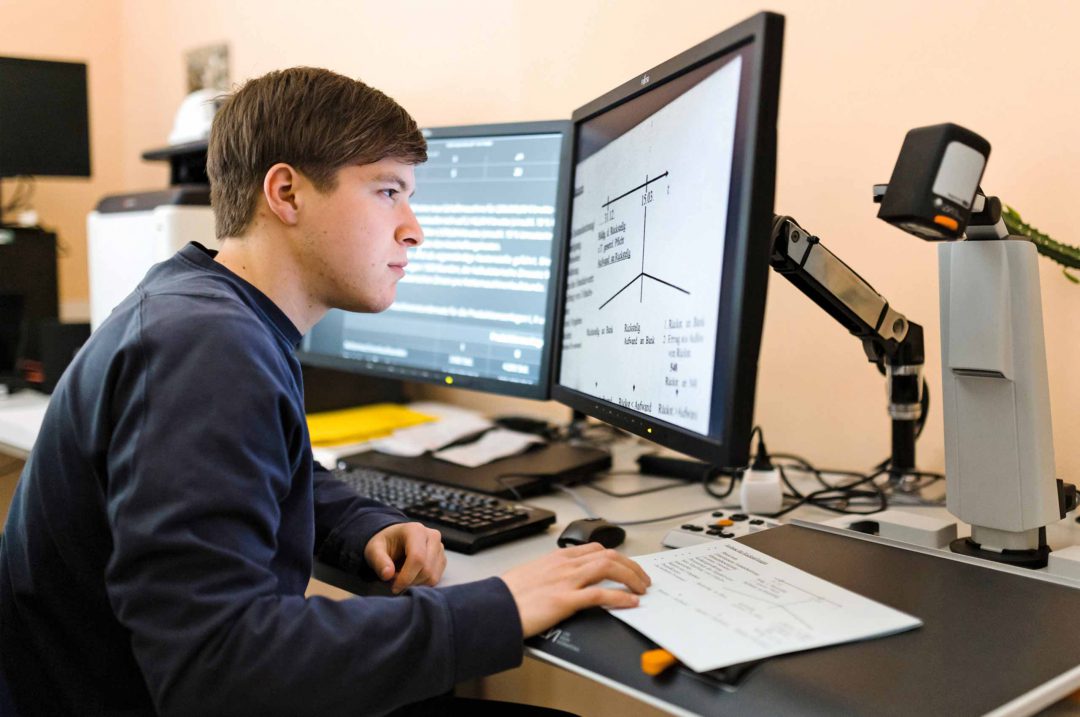 Bildschirme mit enorm vergrößerter Schrift, eine Spezialkamera liest daneben vom Papier ab – so kann Azubi Florin Zapf eigentlich ganz normal arbeiten